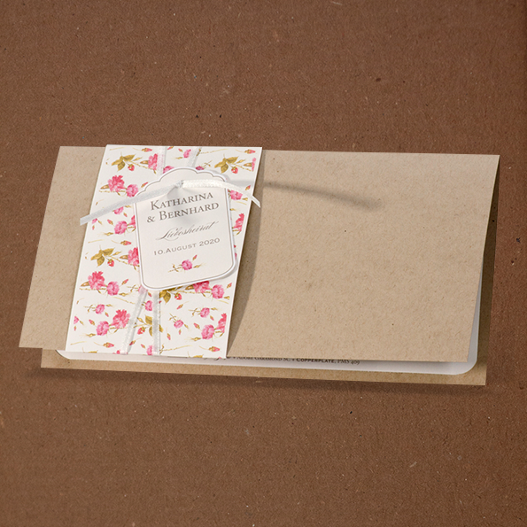 Hochzeitseinladungen Vintage Romantisch – vergrößerte Abbildung Hochzeitseinladung aus braunem Kraftkarton, links umfasst von einer weißen, mit rosa Blümchen bedruckten Banderole, sowie einem weißen Bändchen an welches ein kleines Kärtchen geknüpft ist.