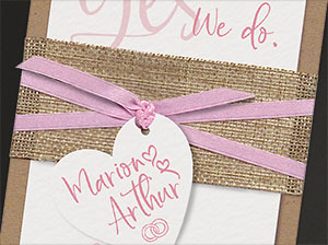 Vergrößerte Abbildung Fertig montierte Einladungskarte. Kraftkarton als Träger, Aquarellkarton mit den Texten, Banderole, Band und angeknüpfter Herzanhänger.