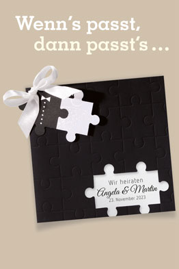 Schwarze Einladungskarte zur Hochzeit, an die mit einer weißen Schleife zwei Puzzleteile angeknüpft sind.